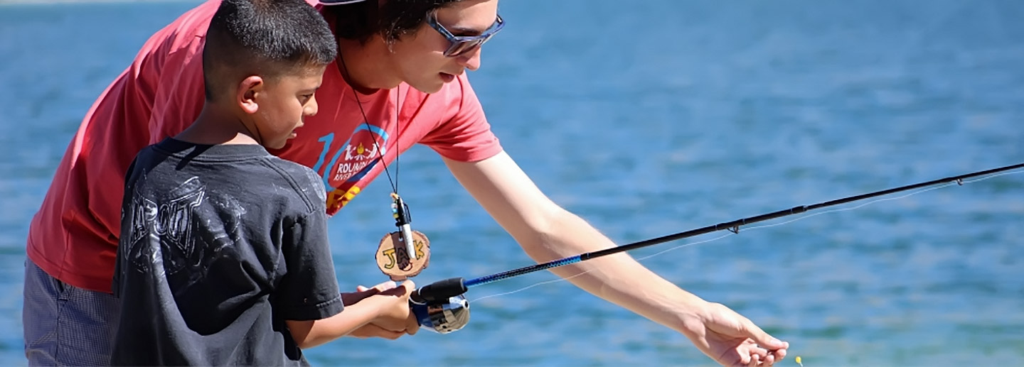 A photo of a volunteer camp counselor teaching a child to fish illustrates volunteer opportunities and summer camp counselor opportunities at Colorado summer camp