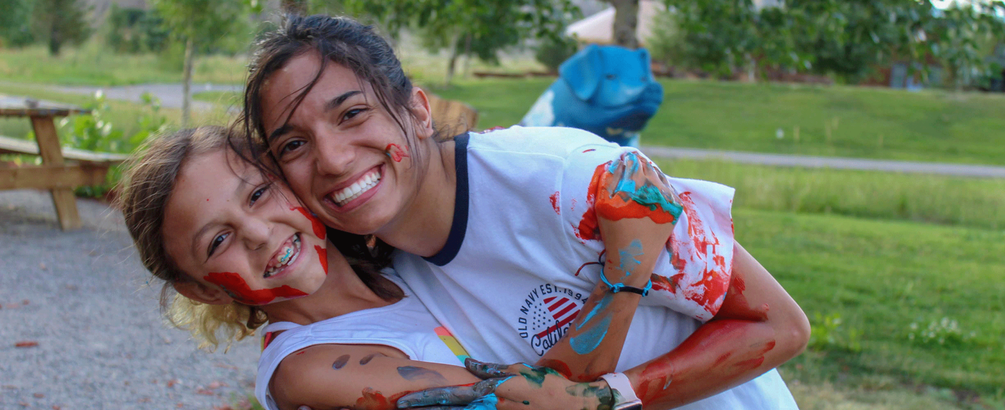 Two campers smiling and embracing after participating in a camp activity that involved orange and blue paint. Apply for camp today.